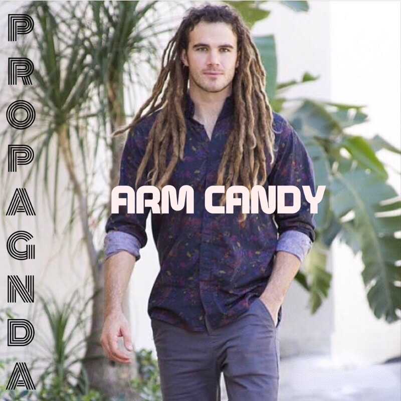 Arm Candy cover art displays an attractive male model with dreadlocks looking confidently toward the viewer in a purple dress shirt with faint flowers. Out of focus palm trees can be made out in the back. 